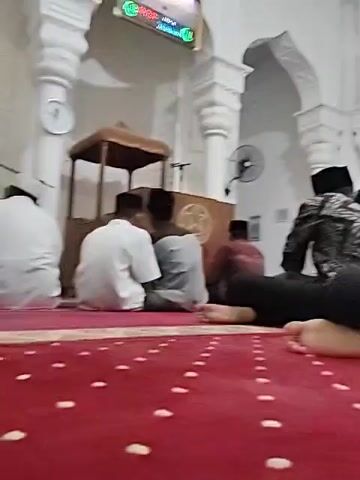 Several men are sitting on a red carpet in a mosque. Most of the men are wearing caps. The mosque is mostly white with columns and arches. There is a wooden pulpit with a star design on it, and a digital clock shows the time. A TV screen displays information in the corner. Some of the men are barefoot.