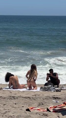 Three people are on a sandy beach with the ocean in the background. A person on the left, wearing a patterned bikini, is leaning forward on their hands and knees. In the center, another person, also in a bikini, is kneeling with their back to the camera, adjusting their hair. To the right, a person in a dark shirt is crouching down and holding a phone, seemingly taking a photo or video. Two towels and a bag are on the sand near the person with the phone. The water has gentle waves.