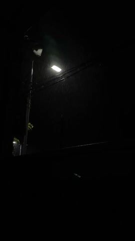 The video depicts a dark, rainy night scene. A street light illuminates falling rain. A power pole is visible with wires running from it. In the distance, another street light can be seen. A dark wall is visible in the foreground. There are no people visible.