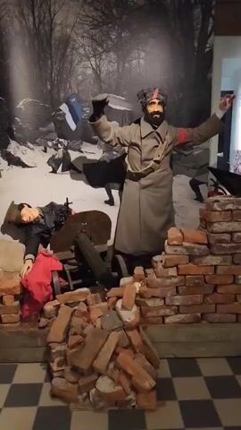 The video shows a museum diorama depicting a scene from a historical conflict. In the foreground, a mannequin of a soldier in uniform lies on the ground next to a machine gun, with a red cloth draped over it. Another mannequin, dressed in a long coat and hat, stands behind a pile of bricks with its arms raised, holding a blue and white flag. The background features a black and white mural of a snowy forest with other figures in the distance. The floor is a black and white checkered pattern.