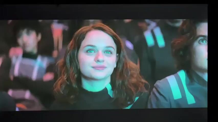 This video appears to be a scene from a science fiction movie or show. The first shot is a close-up of a young woman with brown hair and green eyes looking to her right with a slight smile. She is wearing a black turtleneck. In the background, there are other people dressed in futuristic-looking uniforms, some of whom are out of focus. The second shot shows a woman with long, wavy blonde hair wearing a white strapless dress. She is looking to her left and appears to be speaking or singing, with a dramatic, glowing blue and green background. The scene is lit with neon and atmospheric lighting.