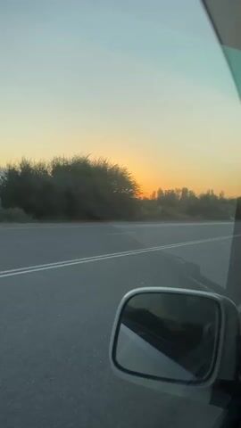 The video shows a sunset viewed from the passenger seat of a moving vehicle. The sun is visible on the horizon, casting a warm glow over the landscape. The road ahead is asphalt with white lines, and trees line the side of the road. A side-view mirror of the car is visible in the lower right corner of the frame. The sky is a gradient of orange, yellow, and blue. The footage appears to be taken during the golden hour.