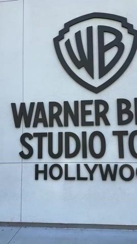 The video shows a close-up, eye-level shot of a white wall with black lettering that reads "WARNER BROS. STUDIO TOUR HOLLYWOOD." Above the lettering is the iconic Warner Bros. shield logo. To the right of the lettering, a dark-colored statue of Wonder Woman is visible. The statue stands on a circular base, and only the lower portion of the statue is in frame. A person is partially visible in the background to the right, standing behind the statue. The lighting suggests it is daytime.