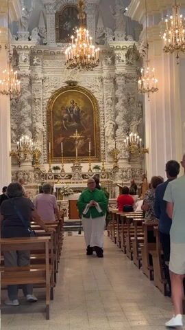 A priest wearing a green vestment over a white robe walks down the aisle of a church towards the altar, which features a large painting of the Virgin Mary and child. He is holding his hands clasped in front of him. Several churchgoers are seated on pews on either side of the aisle, facing the altar. The church interior is ornately decorated with chandeliers and intricate carvings.