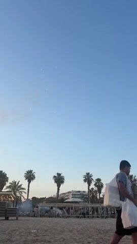 The video captures a beach scene with palm trees in the background. In the sky, fireworks are being set off, with multiple bursts visible against the clear blue sky. Some smoke from the fireworks drifts in the air. Below the fireworks, there is a beach with sand, lounge chairs, and some people. The lighting suggests it is late afternoon or early evening.