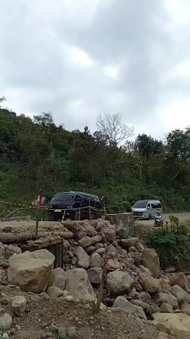The video shows a black van stopped on a rough, makeshift bridge over a rocky area, likely a dried riverbed. The bridge is partially barricaded with yellow and black caution tape.  There's a second, lighter-colored van visible further down the road. The environment is lush with green trees and foliage, giving a forested or rural impression. The sky is overcast with cloudy. A man walks down the road on the left side of the frame.