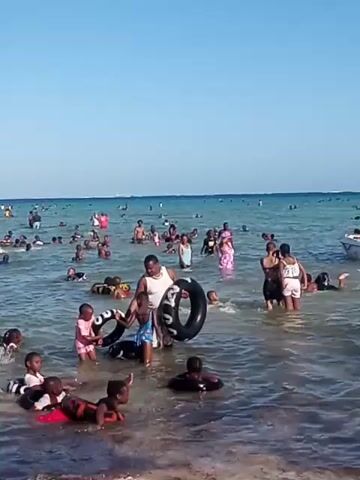 The video shows a crowded beach scene with many people, mainly children, in the water. Most of them are swimming or wading in shallow water near the shore. Some are using inner tubes or flotation devices. An adult male is seen helping a young child with an inner tube. An adult female walks through the water in the foreground. The sky is clear and blue, suggesting a sunny day. The water appears relatively calm. There is a boat visible in the distance.