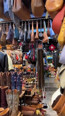 The video shows the interior of a store selling leather goods. Hanging from the ceiling are numerous leather handbags in various colors and styles. Below the handbags are rows of cowboy boots and other leather boots in different colors and designs. A mannequin wearing a leather jacket stands among the boots. A woman is visible squatting in front of a mirror, presumably trying on shoes. Other customers are browsing in the background. The store is well-lit and packed with merchandise.