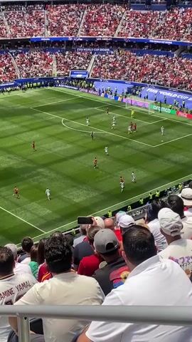 A wide shot from the stands of a packed soccer stadium shows a soccer game in progress. The field is bright green, with white markings. Several players in red and white uniforms are scattered across the field, some running, some standing. In the foreground, a crowd of spectators is visible from behind, all facing the field. They are dressed in casual clothing, and some are wearing hats. The stands are filled with a sea of red and white, indicating a large and engaged audience. The atmosphere appears to be lively, with some spectators raising their hands in the air. The overall scene captures the excitement of a live sporting event.