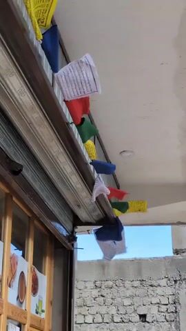 The video shows a close-up, low-angle view of prayer flags strung along a metal awning above a shop. The flags are colorful, with yellow, blue, red, white, and green fabrics, and some have text printed on them. The shop below has a glass front with posters displayed inside. A metal security shutter is partially lowered over the shop. The background consists of a light-colored ceiling and a rough, grey brick wall. The sky is visible through a gap in the awning. The wind appears to be blowing, causing the flags to flutter.