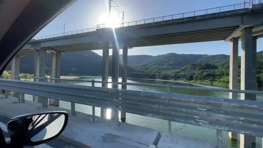 The video shows a view from a car moving across a bridge. The sun is bright and partially obscured by the bridge's structure, casting lens flare across the scene. The bridge has multiple concrete pillars supporting a train track above. Below the bridge is a body of water, likely a lake or reservoir, surrounded by green, rolling hills. The water's surface reflects the sunlight. In the foreground, the car's side mirror is visible on the left side of the frame. The guardrail of the bridge runs along the right side of the view. The weather appears to be clear and sunny.