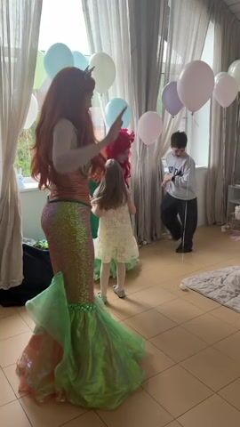 The video captures a lively children's party indoors, featuring a performer dressed as Ariel, the Little Mermaid, in a glittering mermaid tail costume. Beside her is another person costumed as a red-haired mermaid, possibly also Ariel. Around them, several young children, including a girl in a white dress, dance and interact with the costumed figures. 

The room is decorated with balloons in pastel shades, and a large window with sheer curtains provides natural light. In the background, other children are jumping and dancing on a large mattress laid on the floor, near toys, a baby stroller, and a bookshelf. The scene is cheerful, with the children seemingly enjoying the entertainment and music.