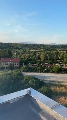 The video pans across a landscape from a high vantage point. In the foreground, a white ledge or railing is visible, with some debris or wear on its surface. The scene transitions to reveal a rural or semi-rural area with scattered houses and buildings amidst green vegetation and rolling hills. A single-story building with a red roof and light-colored walls is prominent in the mid-ground. Further into the distance, the landscape becomes more mountainous. The sky is a clear blue with scattered white clouds, and the sun is low in the sky, casting a warm glow and creating lens flares in some shots. Towards the end of the video, the view shifts to show a cluster of apartment buildings, suggesting a transition into a more urban or suburban area. The sun is brighter and higher in the sky in these latter shots, with pronounced sunburst effects. The overall impression is a picturesque view of a landscape at either sunrise or sunset.
