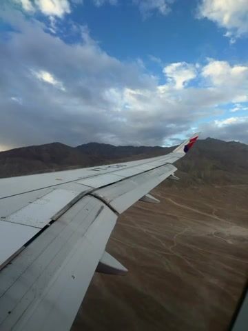 This video shows a view from an airplane window as the aircraft flies over a desert landscape with mountains in the background. The airplane wing is visible in the foreground, and its flaps are extended. The sky is partly cloudy, with patches of blue sky visible between the clouds. The sun appears to be low on the horizon, casting a warm light on the landscape. The video captures the movement of the airplane as it flies over the terrain, with the landscape appearing to move from right to left. The overall impression is one of a peaceful and scenic flight.