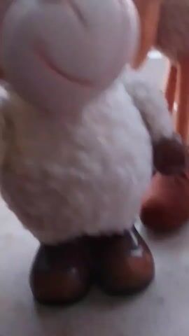 A close-up video shows two plush sheep toys on a windowsill. The sheep in the foreground is white with brown hooves and light brown ears. It has a pink nose and blue eyes and is facing the camera. The sheep in the background is a pinkish-brown color and is standing on long, thin orange legs with brown hooves. It has a green and yellow flower on its head. The sheep are positioned near a window with a blurred outdoor scene visible through it.