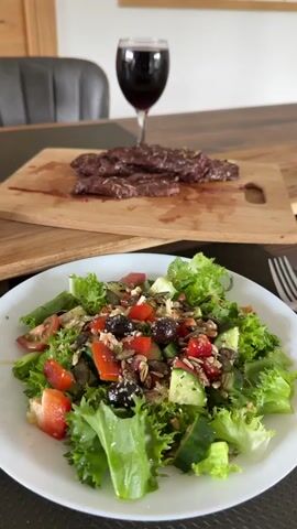 A close-up shot shows a white plate with a salad, featuring lettuce, tomatoes, cucumber, olives, seeds, and possibly cheese. Behind the plate is a wooden cutting board with sliced steak and a wine glass filled with red wine. The setting suggests a meal is ready to be enjoyed. The camera focuses on the details of the salad and steak, presenting an appealing view of the food arrangement.