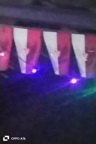 A series of flags, alternating red and white with a symbol in white on the red portion, are strung horizontally and appear to be illuminated by colored lights, including purple and green. The flags are hanging from a wooden structure, possibly a roof or awning. The background is dark, suggesting it is nighttime or a dimly lit area. A watermark at the bottom left corner reads "OPPO A76".