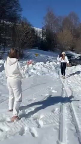 Two women engage in a snowball fight in a snow-covered landscape under a clear blue sky. The woman on the left is wearing a white sweatsuit, while the woman on the right is dressed in black pants and a white jacket. They are standing apart in a snowy area with tire tracks. Behind them are trees and parked cars. The woman in white throws a snowball toward the woman in black.