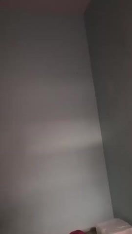 The video pans upwards revealing a ceiling fan in motion. The walls are painted a light blue and there is a corner where two walls meet. On the walls, there is a clock and what appears to be a calendar. There are also shelves on the left side of the frame with various items on them. The lighting in the room is dim.