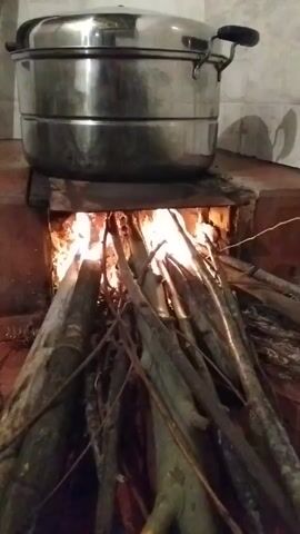 This video shows a close-up of a steamer pot sitting atop a traditional wood-fired stove. The fire within the stove burns brightly with visible flames and glowing embers, casting a warm light on the surrounding materials. A collection of dry branches and logs are piled around the opening of the stove, ready to be added to the fire. The steamer pot, made of stainless steel, has two tiers and two handles on its lid. It is positioned directly over the heat source. The stove itself appears to be constructed from brick or clay, with a metal plate serving as a platform for the pot. The background is dimly lit, with hints of tiled walls. The overall scene suggests a rustic cooking method, possibly in a rural or outdoor setting. No people are visible in the frame. The activity depicted is the heating of water or food within the steamer pot using a wood fire. The image is well-lit from the fire, creating a high-contrast scene. There is no visible text in the video.