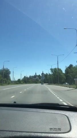 The video is a first-person view from inside a car driving on a paved road. The sky is clear and blue. On the left side of the road, there are trees and a few buildings, including a white church with a dark dome in the distance. On the right side of the road, there are more trees and streetlights. Several cars are visible on the road ahead, moving in the same direction as the camera. The dashboard of the car is visible in the foreground, with an "SRS AIRBAG" label.