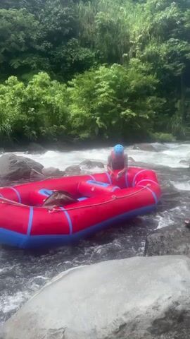 A person in a blue helmet and a life vest is in a red inflatable raft on a river. They are holding a white pump with a gray handle and appear to be inflating the raft. The raft has blue trim. A brown bag is in the raft. The river has rapids and rocks. Lush green foliage is in the background, suggesting a tropical environment. The scene appears to be set during the day. The person's facial expression is neutral or serious. The location is Selat, Indonesia.