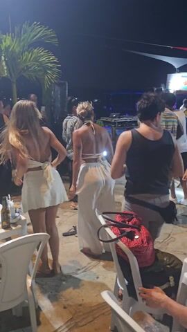 At night, people dance at an outdoor venue. Two women in white outfits are nearest the camera, along with others dancing. Many people are in the background, enjoying the party. Tables and chairs are placed nearby, along with red and black bags on one chair. A palm tree stands off to the left.