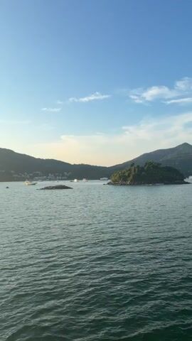 The video captures a scenic view of a bay with calm water, framed by lush green hills under a clear blue sky with scattered clouds. Several boats are visible in the distance, either anchored or gently moving across the water. The sun is setting, casting a warm glow and creating a serene atmosphere. The perspective suggests the footage is being filmed from a boat, with a portion of the boat's seating and canopy visible in the foreground, particularly in the latter half of the video. The light shifts from bright daylight to a softer, golden hour hue as the sun descends behind the hills.