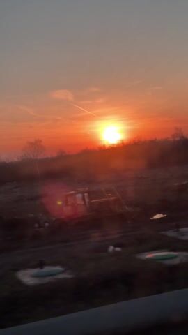 The video shows a sunrise over a construction site. The sun is bright orange, and there is a contrail in the sky. The foreground is dark, but there are several pieces of construction equipment visible in silhouette. The camera moves slightly during the recording, and a window with condensation is visible at the end.