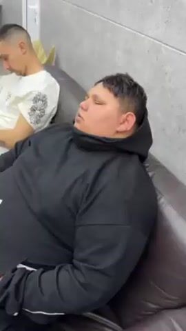 The video shows a young man wearing a black hooded sweatshirt, asleep on a brown leather couch. He is on his side, facing toward the camera. Another person sits beside him, but is only partially visible.  The room has light gray walls.