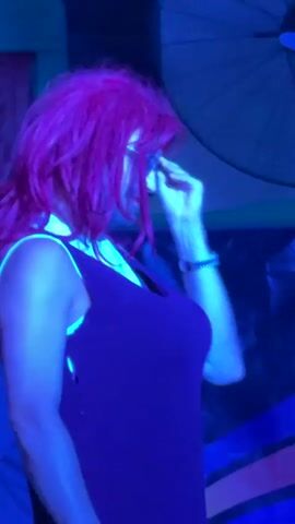 A drag performer with bright pink hair and sunglasses, wearing a purple dress over a white shirt, holds a microphone and gestures with their right hand. The performer is on a stage with red curtains in the background. Another performer, also with dark hair and sunglasses, is visible to the left of the main performer. The stage is lit with purple and blue lights. The video appears to be a recording of a live performance.