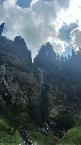 The video captures a serene natural scene featuring a towering, rocky cliff face with a thin waterfall cascading down its side. Lush green vegetation covers the lower slopes of the cliff, and a pathway with stone steps leads upwards towards the waterfall. Several people are seen ascending the steps, suggesting a pilgrimage or tourist destination. At the base of the waterfall, a small temple or shrine is visible, adorned with flags and offerings. The sky above is partly cloudy, with rays of sunlight breaking through, illuminating the scene and highlighting the mist from the waterfall. The overall atmosphere is one of natural beauty and spiritual tranquility.