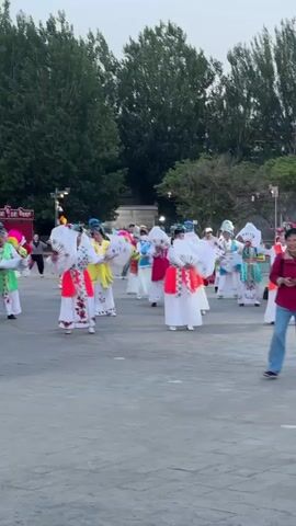 A group of women dressed in colorful traditional Chinese outfits perform a dance with white fans on a grey brick-paved area. There are trees and lampposts in the background. One woman, dressed in jeans and a red shirt, walks in front of the group, filming them with her phone. The dancers are positioned in rows and moving forward.