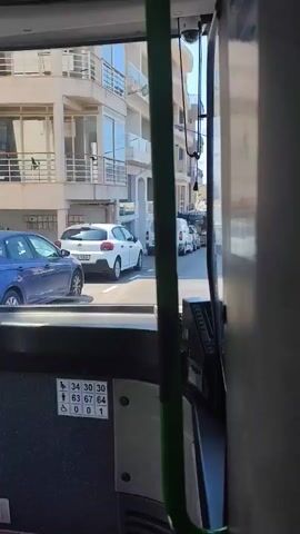 The video appears to be filmed from inside a bus, looking out the front window. The view is of a street scene, with buildings lining both sides. Several cars and vans are parked along the street, and a few people are visible. The bus seems to be in motion, as the perspective shifts slightly throughout the recording. There are also some signs visible inside the bus, including what appear to be seating priority indicators.