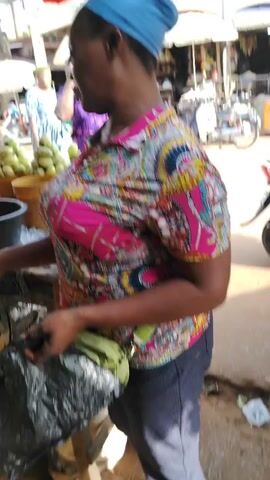 Here is a concise description of the video:

The video shows a bustling marketplace scene, possibly in Africa. The camera focuses on a person wearing a brightly colored shirt and a blue head covering. The scene then shifts to a stall overflowing with round, green vegetables in buckets and containers. A woman wearing a hat is seen pushing a wheelbarrow loaded with groceries down the street. The general environment appears to be a lively, outdoor market area.