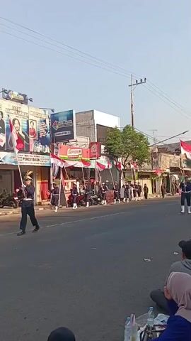The video shows a parade on a paved street. A group of young people in uniform, wearing dark blue jackets, white pants, and white shoes, march in formation carrying Indonesian flags. They are walking from left to right across the frame. On the left side of the street, there are buildings with signs and posters, including one with a picture of a person. On the right side of the street, there are spectators sitting on the ground, also facing the parade. The sky is clear and bright, suggesting it is daytime. There are also some parked motorcycles visible on the left side of the street. The overall scene depicts a public event, likely a celebration or commemoration.
