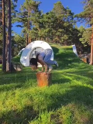 A person is performing a yoga pose called Bakasana (Crane Pose) on a tree stump in a grassy, wooded area. The person is balancing on their hands with their knees tucked into their chest. Behind the person is a unique, round, white structure built into a grassy hill, resembling a hobbit house. Tall pine trees surround the area, and the sky is clear blue. The person's hair is dark and falls forward as they hold the pose. The scene appears to be taking place outdoors on a sunny day.