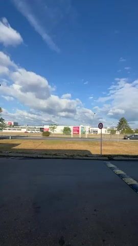 The video shows an outdoor scene with a blue sky filled with white clouds. In the foreground, there is a speed bump on an asphalt road. Behind the speed bump, there is a grassy area and a road. On the other side of the road, there is a row of buildings with signs, including one that says "OMV". There are also trees and several cars visible. The scene appears to be a gas station or a commercial area. The camera pans from left to right, showing different parts of the area.