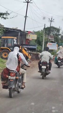 A group of people are riding motorcycles on a paved road. The motorcyclist in the foreground is carrying a woman behind him. The woman is wearing a light-colored headscarf and a red skirt. The motorcyclist is wearing a light-colored shirt and tan pants. Another motorcycle is visible in the background, ridden by a man in a plaid shirt. A white car is parked on the side of the road. A yellow excavator is visible behind a building on the left side of the image. The sky is overcast.