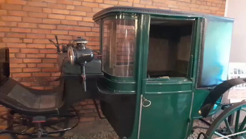 The video shows a close-up, side-view, medium shot of a dark green carriage with black leather accents, parked indoors. The carriage has a glass-enclosed passenger compartment with a dark interior. To the left of the passenger compartment is a driver's seat. A black kerosene lamp is mounted on the left side of the carriage, in front of the passenger compartment. The carriage is positioned in front of a brick wall. The floor appears to be covered with gravel or small stones. There are no people or animals visible in the video. The lighting suggests an indoor setting, possibly a museum exhibit.