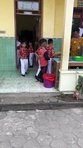 A group of young school children, dressed in red and white uniforms with some wearing white headscarves, are lined up outside a building entrance, possibly for a meal or activity. An adult male, wearing a patterned shirt, stands behind a table laden with stacked metal trays, serving or preparing to serve something. A green bowl is visible on the table. A purple trash can with a red liner is situated near the children. The environment appears to be an outdoor school setting, with a tiled pathway and yellow walls of the building.