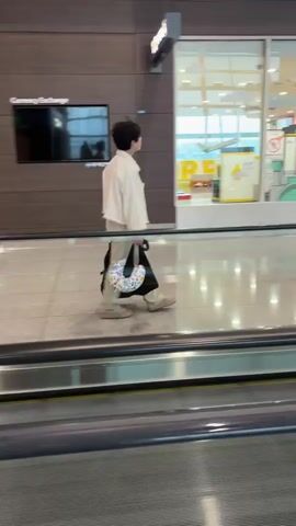 The video captures a wide shot of a person walking on a moving walkway, likely in an airport or a large indoor public space. The subject, seen from the side, is wearing a light-colored, loose-fitting jacket over pants, and carrying a patterned neck pillow bag over their shoulder. They have short, dark hair and are looking forward.

To the right of the subject, another person is walking in the same direction. This second individual is wearing a dark t-shirt, carrying a tote bag, and appears to be looking at their phone.

In the background, there is a large storefront with glass windows. Visible signage on the store reads "KIDS AIRPORT" in English and "키즈포트" in Korean. The interior of the store appears bright and colorful, with displays of what might be toys or play structures. A security camera is also visible mounted on the wall above the storefront.

The floor is polished and reflective, showing the lights from above. The moving walkway itself is prominently in the foreground, with its metallic handrails and textured surface. The overall lighting suggests it is daytime, and the indoor setting indicates it is not weather-dependent. The mood appears neutral, with people engaged in typical travel or transit activities.