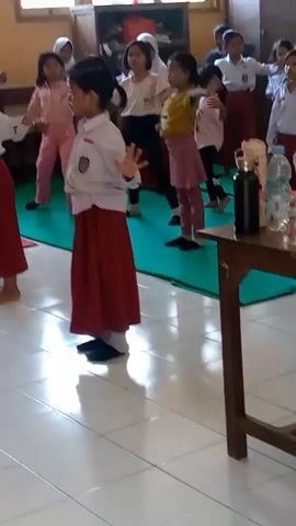 A group of young girls in uniform are engaged in a coordinated physical activity, likely a dance or exercise routine, in a classroom. The girls are wearing white shirts with badges and red skirts, and some have headscarves. They are arranged in rows and appear to be following instructions or a demonstration. The environment is a classroom with tiled floors, wooden furniture, and windows. A fan is visible in the foreground. The lighting suggests it is daytime. The actions of the girls are synchronized and involve arm movements and body poses.