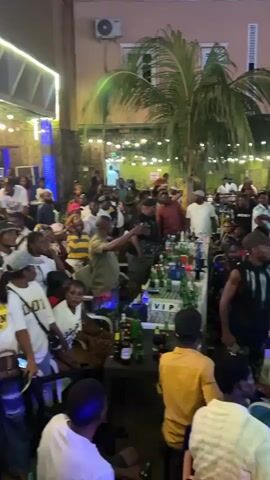 The video shows a crowded outdoor event at night. People are gathered around tables, some of which are laden with bottles of drinks. There are palm trees and string lights decorating the area. The mood appears festive, with many individuals dressed in casual attire. The camera pans across the scene, capturing the large number of attendees and the lively atmosphere of the gathering. There are no overt signs of violence or distress.