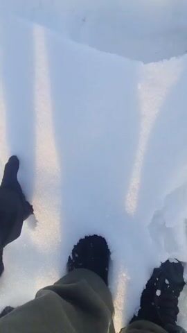 The video begins with a close-up of a gloved hand holding a snowball, standing in snowy ground with bootprints visible. The scene then shifts to a man dressed in a black jacket and light-colored pants, walking away from the camera through the snow-covered ground. He is wearing a face mask and walks past a pile of rocks and debris. In the background, there is a building, a car, a tree, and a partially visible blue sky. The overall environment appears to be on the edge of a residential area.