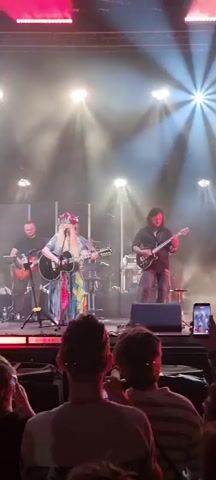 The video shows a band performing on stage in front of an audience. The lead singer, a woman with long blonde hair wearing a colorful dress and a floral headdress, plays an acoustic guitar and sings. To her right, a man with long dark hair plays an electric guitar. To her left, a man with short dark hair plays a bass guitar. Behind the lead singer, a drummer can be seen. The stage is lit with bright spotlights, and there is a moderate amount of haze in the air. The audience members are mostly visible from the back of their heads and shoulders. One audience member in the foreground is holding up a phone to record the performance. The overall scene suggests a live music concert.