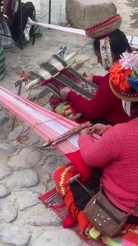 Two indigenous women are sitting on the ground weaving fabric. They wear traditional clothing, including colorful hats. One woman wears a red sweater and has a brown purse slung over her shoulder. The other wears a maroon sweater. They are working on looms set up on the ground. One loom is strung with red yarn, and the other is strung with striped green, purple, and white yarn. The ground is made of cobblestones. In the background are various items including a backpack, a metal stand, and a white bucket.