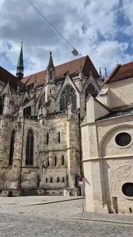 A video captures a time-lapse of a large stone cathedral. In the foreground, a cobblestone street is visible, with a few people walking by. The cathedral features intricate architecture with spires and arched windows, and some scaffolding is present on one of the towers, suggesting ongoing construction or maintenance. The sky above is cloudy, with patches of blue sky peeking through. The camera appears to be stationary.