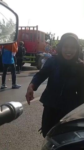 The video captures a street scene with several people and vehicles. In the foreground, a motorcycle's handlebars and side mirror are visible, suggesting the footage is being filmed from a motorcycle. To the left, a person wearing a blue denim jacket and a hijab is looking and smiling towards the camera. In the background, there is a red truck, several tents or makeshift structures, and a group of people. Two individuals in elaborate costumes, possibly dancers, are positioned in the middle of the road, with other people observing them. One person is riding a motorcycle with boxes and a patterned bag strapped to the back. The road appears to be paved, and the environment suggests an outdoor daytime setting, possibly a gathering or event on the roadside.