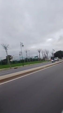 The video shows a gray Volkswagen Touareg driving on a paved road under a cloudy sky. The car is seen from the side and rear, with a brief view of the front at the beginning. There is a strip of greenery and a concrete median between the lane the Touareg is in and the opposing lane. Another car, a silver sedan, drives in the opposite direction. Bare trees and some with leaves are visible along the side of the road, along with streetlights. A soccer field can be seen beyond the trees. The video appears to have been taken from a moving vehicle.